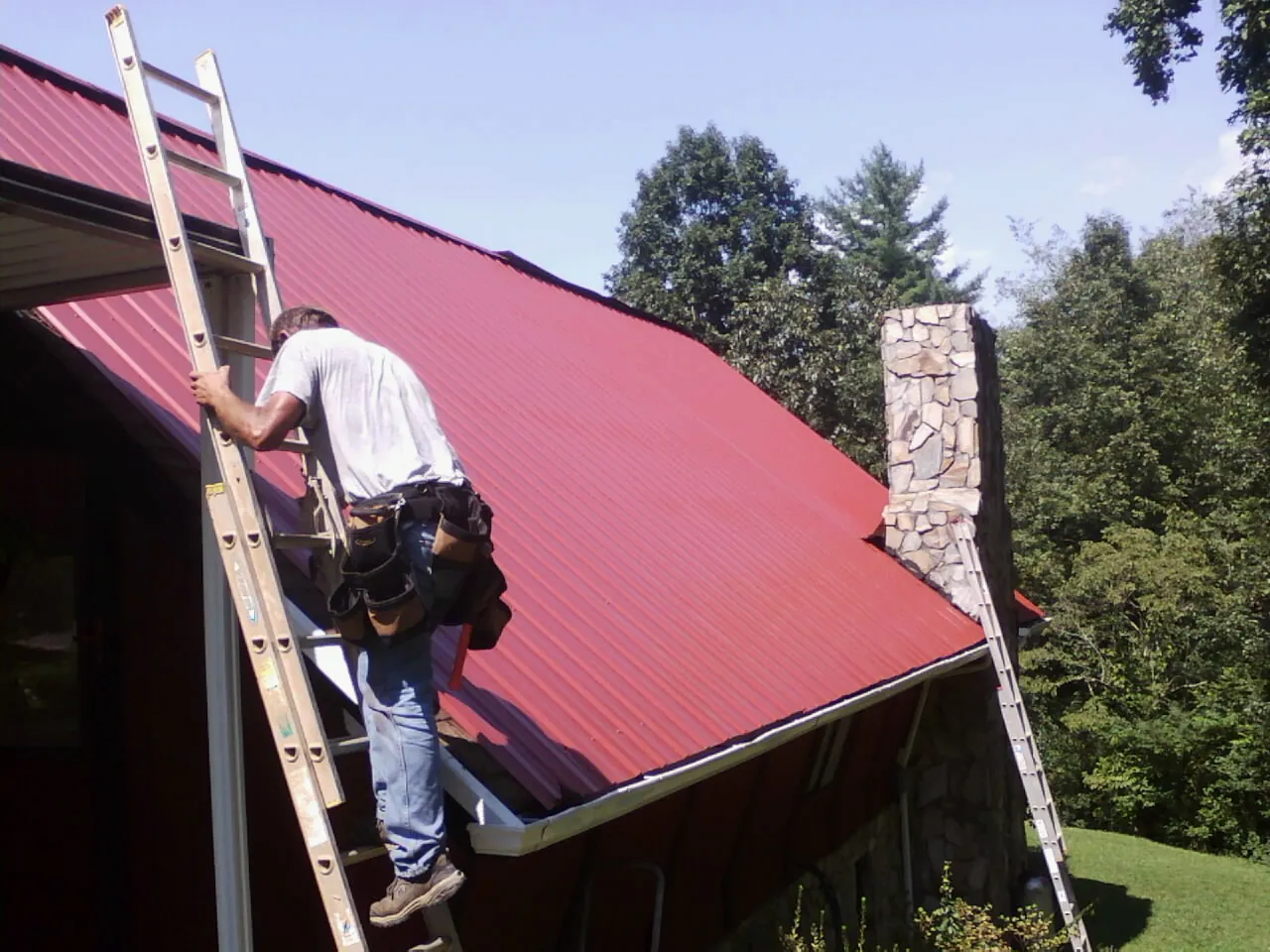 Roofer on a ladder beside a red metal roof and stone chimney — Soffit & Fascia Repair by Roofing Brothers in Shelbyville